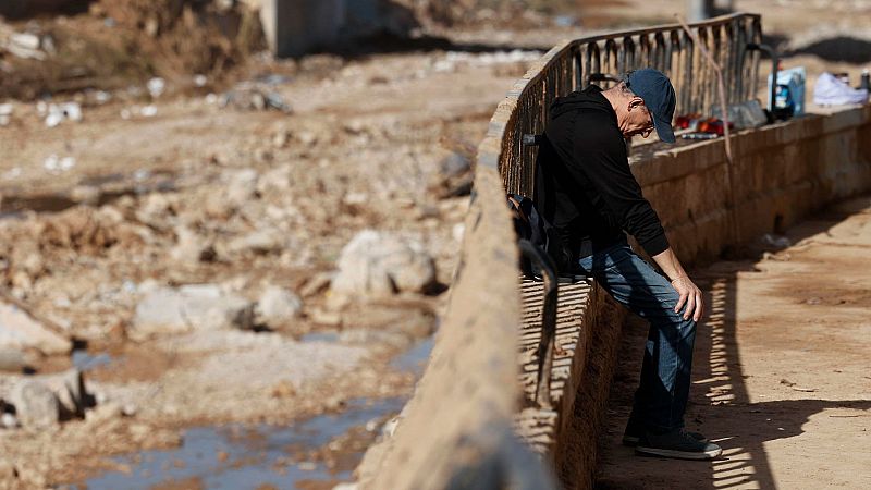 Hombre sentado, contemplando los daños causados por la DANA: barro, escombros y devastación.  El contexto de la noticia sobre la prohibición de subidas de precios añade significado.