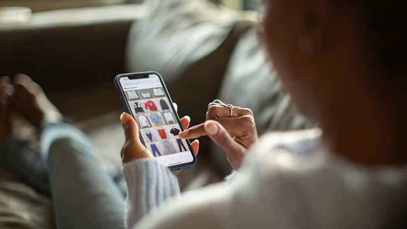 Mujer comprando ropa online en su sofá. Lleva un jersey claro y vaqueros, usando su smartphone.