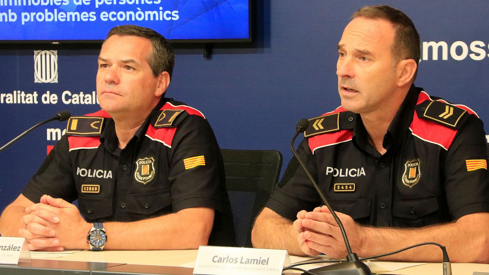 Rueda de prensa de dos Mossos d'Esquadra, Miquel Àngel González (13449) y Carlos Lamiel (5454), con micrófonos y logo de la Generalitat de Catalunya al fondo.