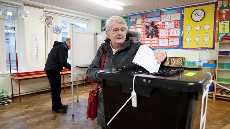 Mujer votando en el colegio electoral St Anthony Boys National School, Irlanda. Lleva abrigo gris, bolso rojo y deposita un papel en una urna negra.