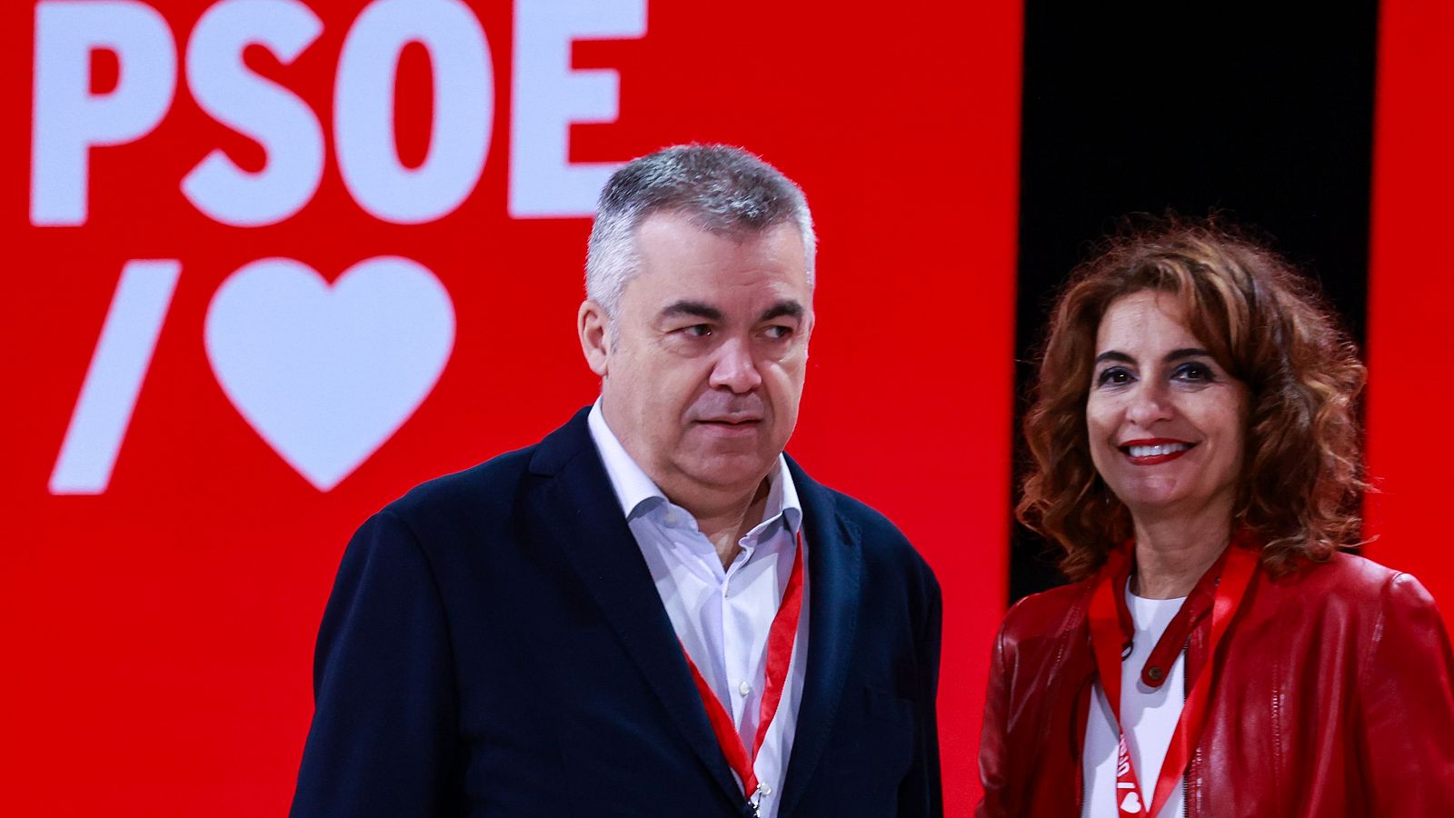 María Jesús Montero y Santos Cerdán del PSOE en un acto del partido. Ambos llevan acreditaciones rojas y posan ante un fondo rojo con el logo del PSOE.