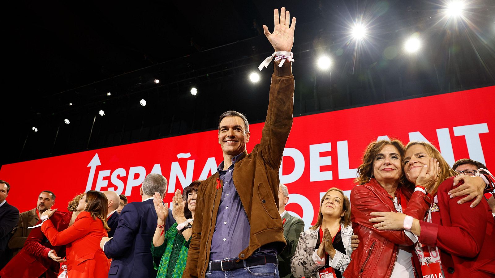 Pedro Sánchez, en el 41 Congreso del PSOE en Sevilla, saluda a la multitud.  Viste chaqueta marrón y camisa azul; detrás, un cartel rojo dice "España Delante".