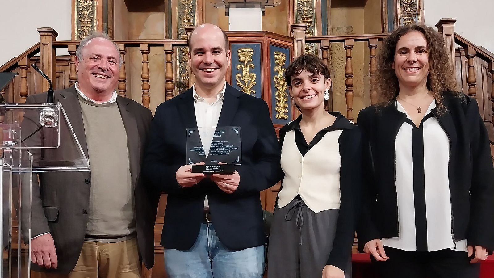 Cuatro personas posan en un auditorio; un hombre sostiene una placa conmemorativa. Evento de reconocimiento.