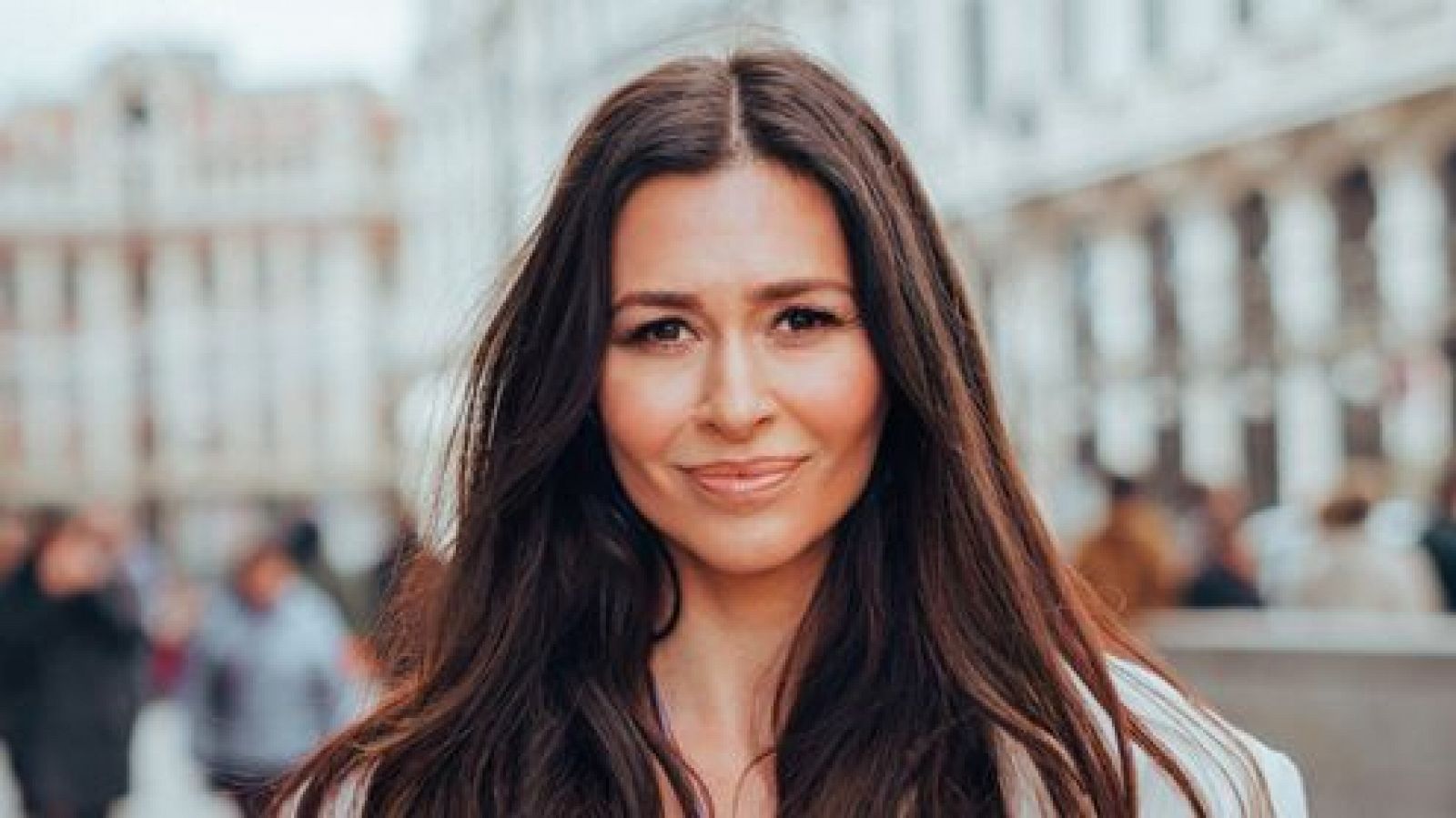 Mujer de mediana edad con cabello castaño, ojos oscuros y una leve sonrisa, vestida con ropa clara, en un entorno urbano.  Texto adicional: "Orquesta y Coro de RTVE".