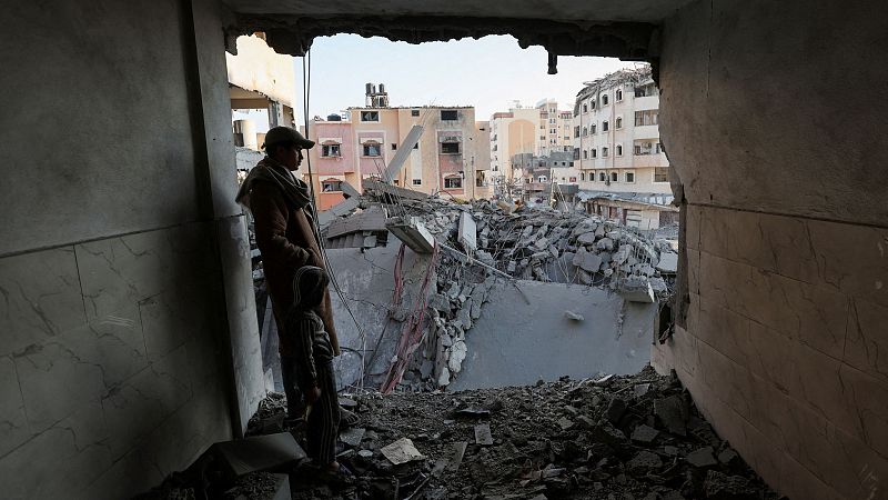 Hombre en edificio dañado de Gaza observa la destrucción.  Viste ropa informal, incluyendo gorra y bufanda.  Escena de devastación con escombros y edificios en el fondo.