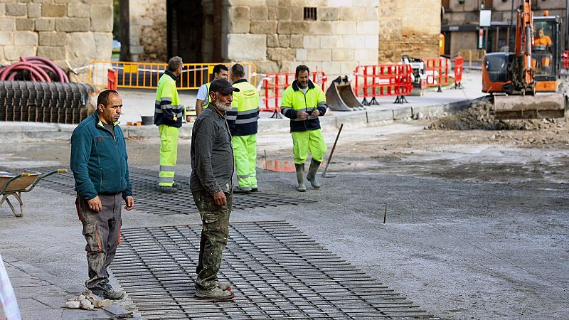 Obreros trabajando en una obra de pavimentación con malla metálica, vallas, excavadora y herramientas.  Se observa actividad constructiva.
