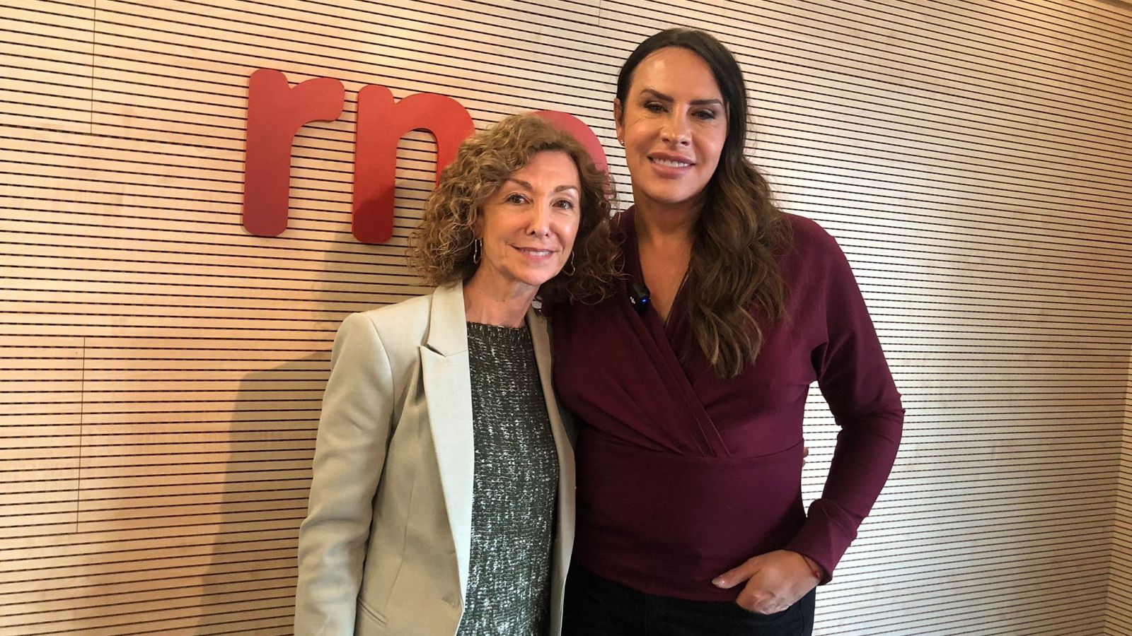 Dos mujeres, una con blusa granate y otra con chaqueta beige, posan sonrientes junto al logo de RNE.  Una de ellas es Karla Sofía.