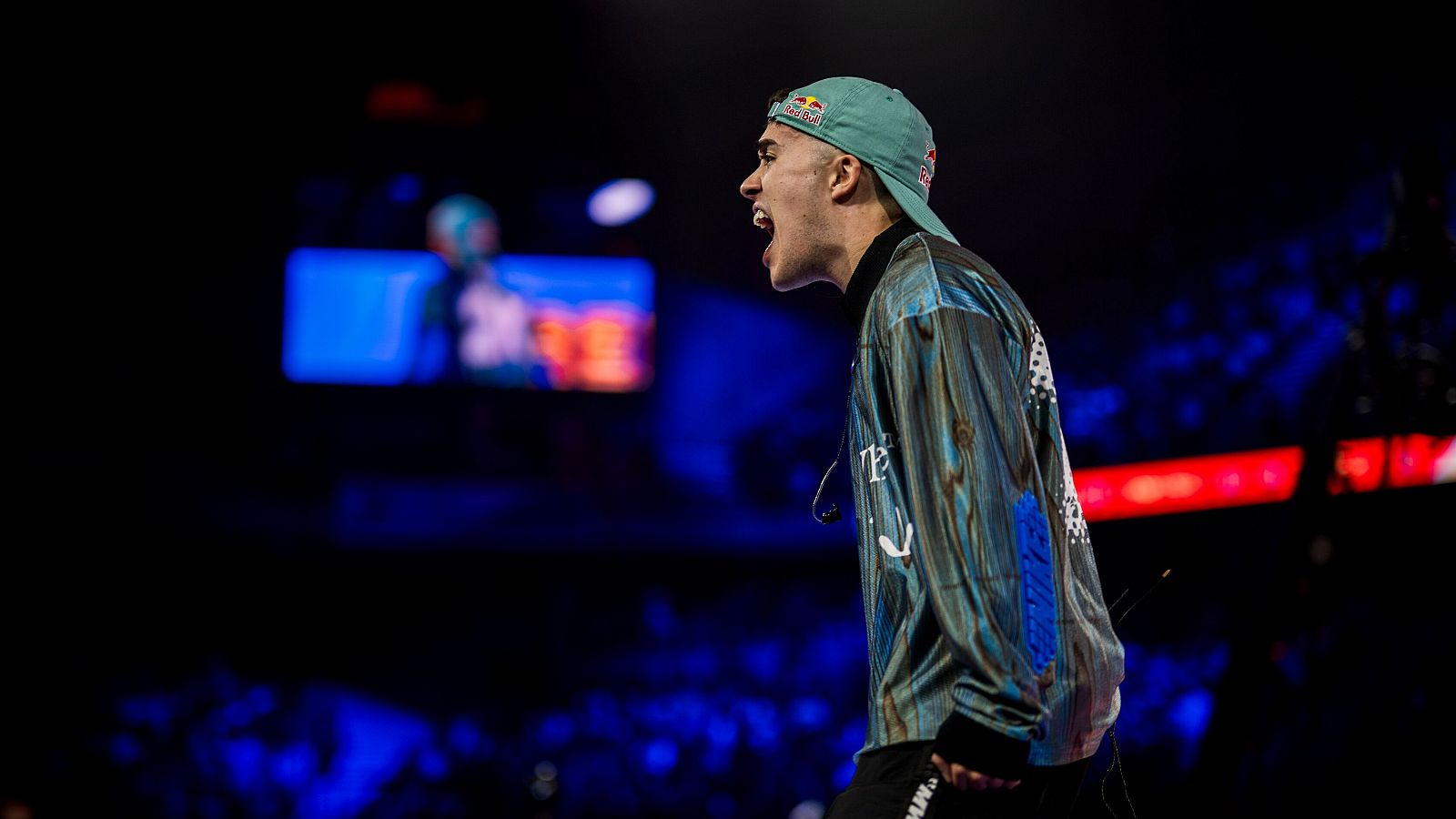 Rapero con gorra y chaqueta actuando en un escenario con luces azules, micrófono cercano y público difuminado al fondo.  Imagen de alta energía.