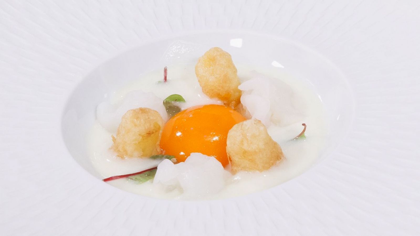 Elegante plato con yema de huevo, esferas blancas y patatas doradas sobre base blanca. Presentación radial, propia de alta cocina.