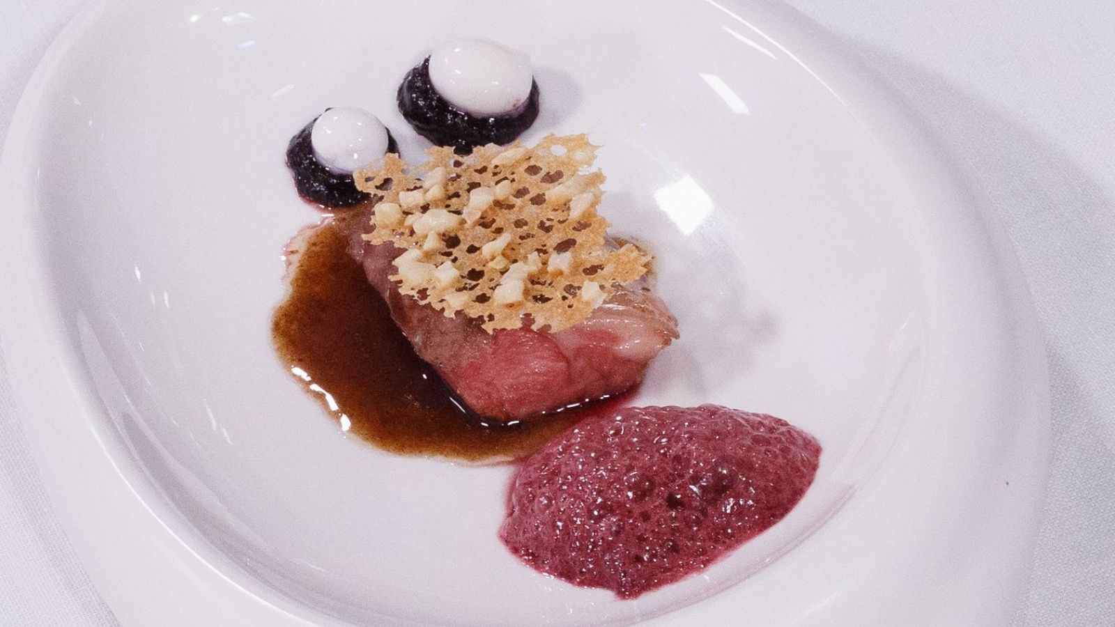 Cordero asado, rosado por dentro, con salsa, puré de cerezas y crujiente con almendras. Presentación elegante en plato blanco.