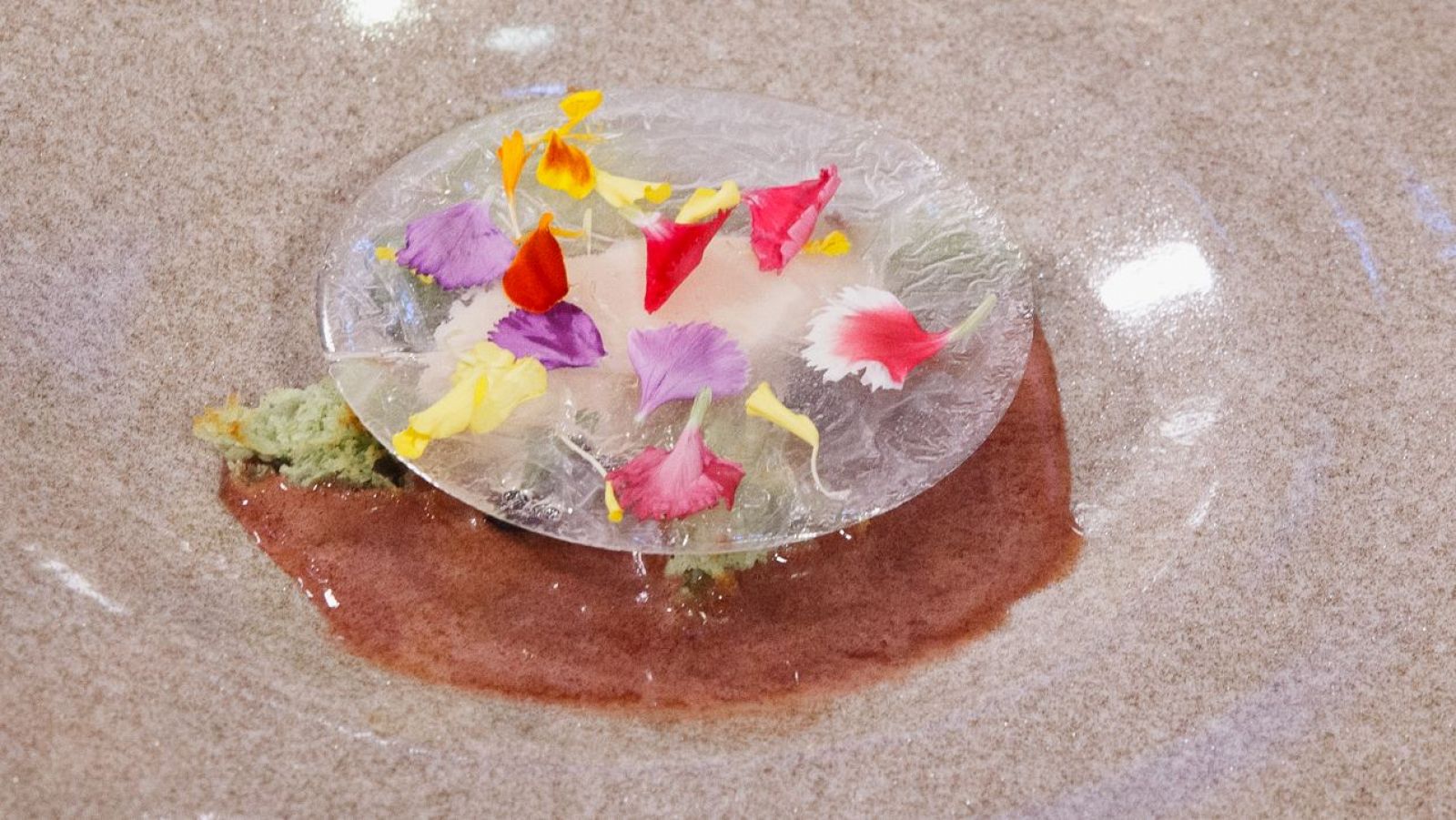 Helado de judía blanca con pétalos y guarnición verde sobre sopa de cereza. Presentación atractiva en un plato de textura rugosa.