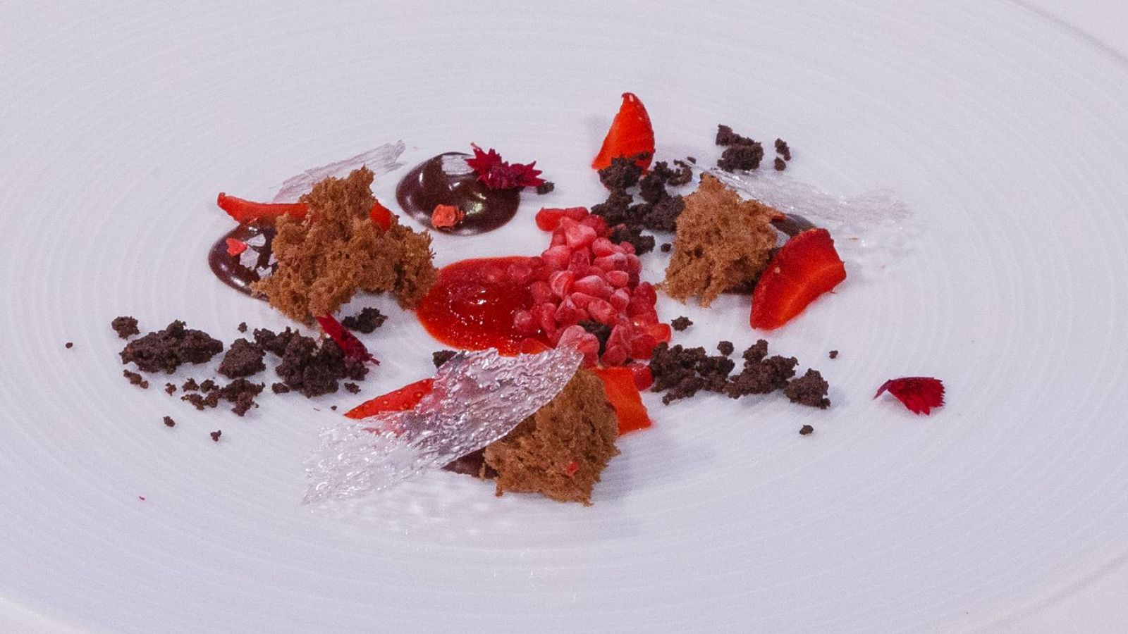 Atractivo postre con fresas, frambuesas y chocolate; bizcocho, salsa roja y láminas transparentes decoran el plato blanco.