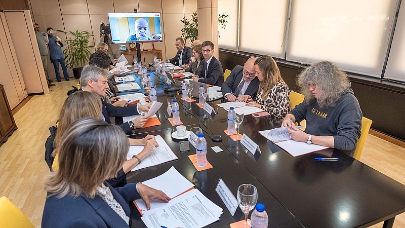 Miembros de un consejo sentados alrededor de una mesa, revisando documentos. Videoconferencia en una pantalla al fondo.