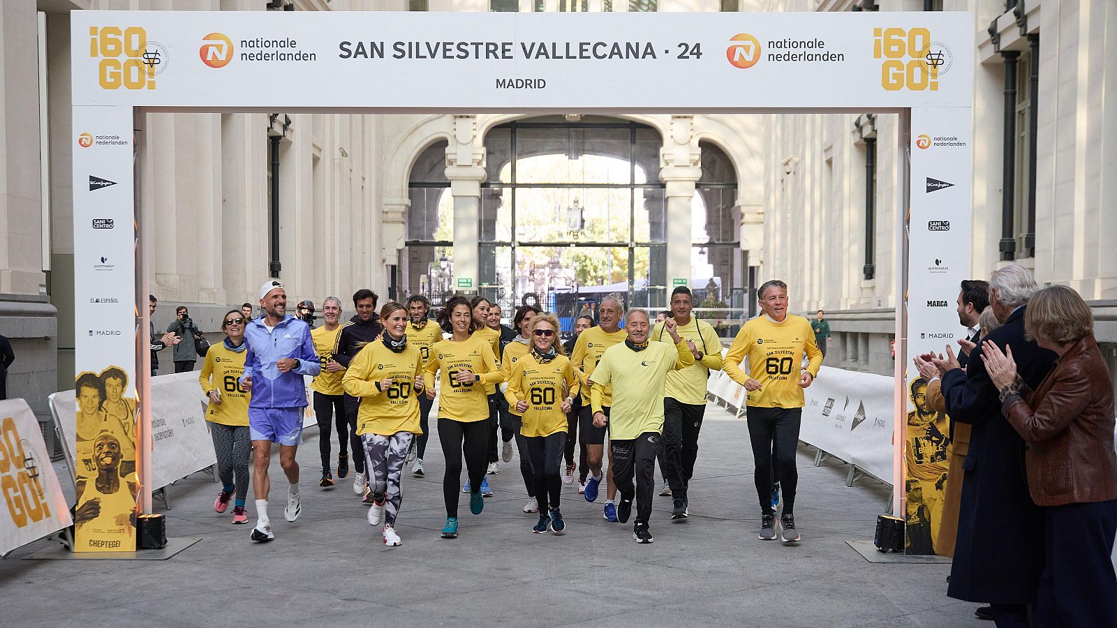 Corredores con camisetas amarillas y el logo "60 GO!" llegan a la meta de la San Silvestre Vallecana, patrocinada por Nationale Nederlanden.  Público aplaude.