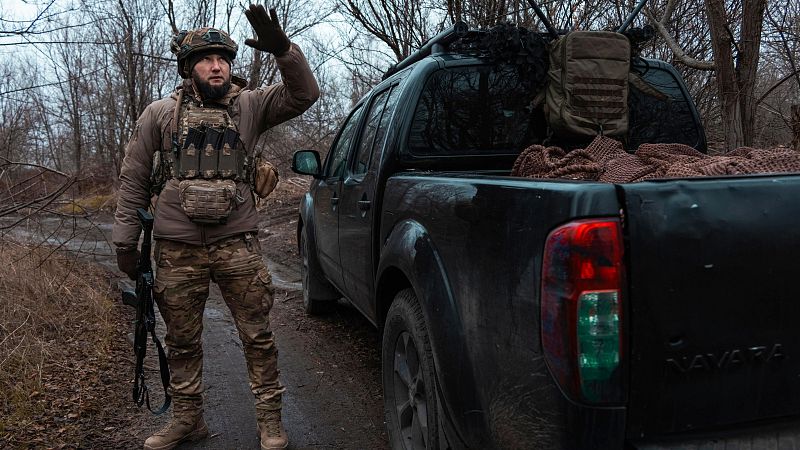 Soldado ucraniano con uniforme, casco y chaleco antibalas, junto a una camioneta pick-up camuflada, hace una señal con la mano mientras porta un rifle.