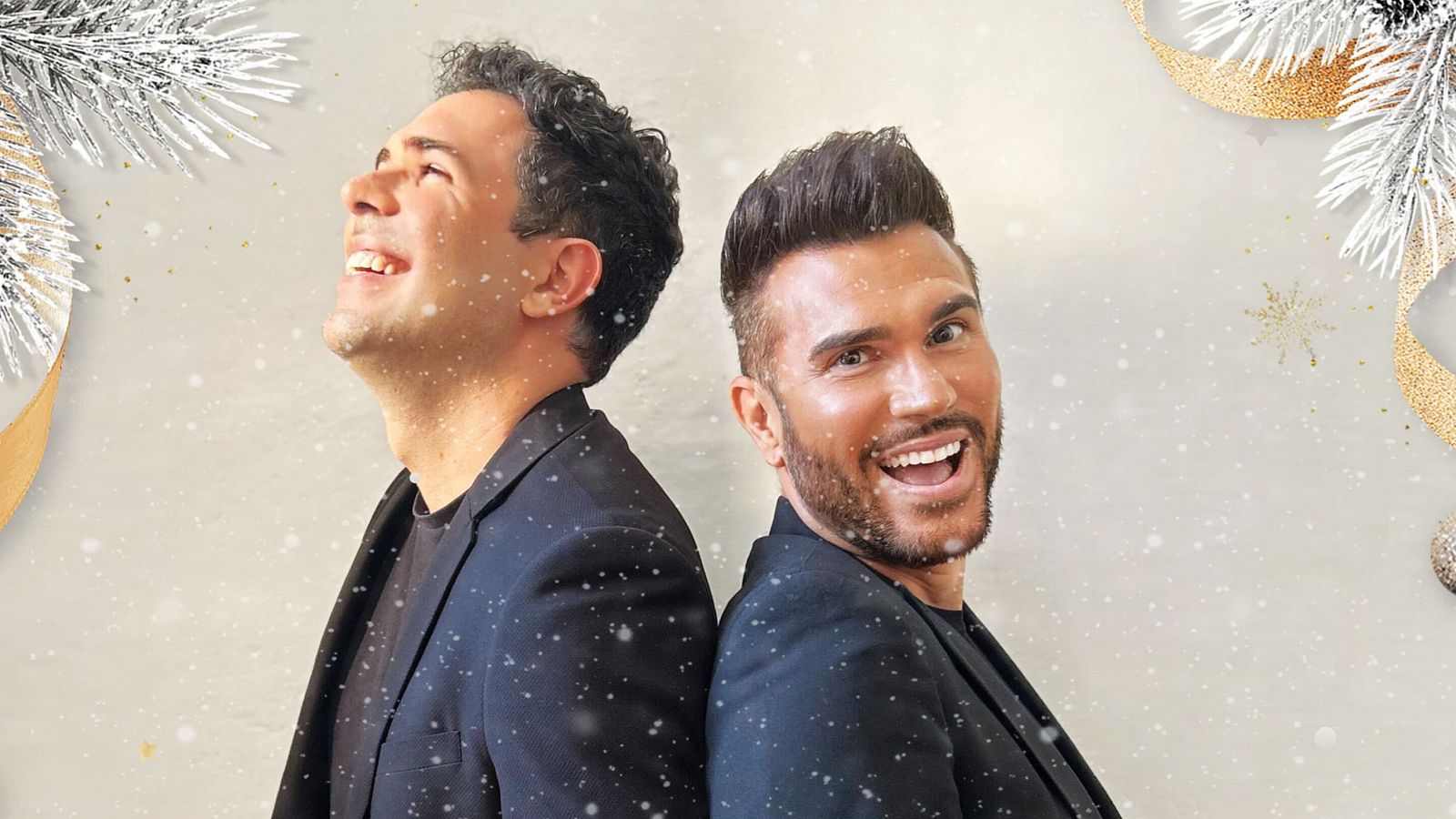 Dos hombres sonrientes, con americanas oscuras, posan espalda con espalda en un ambiente navideño con nieve.  Concierto navideño.