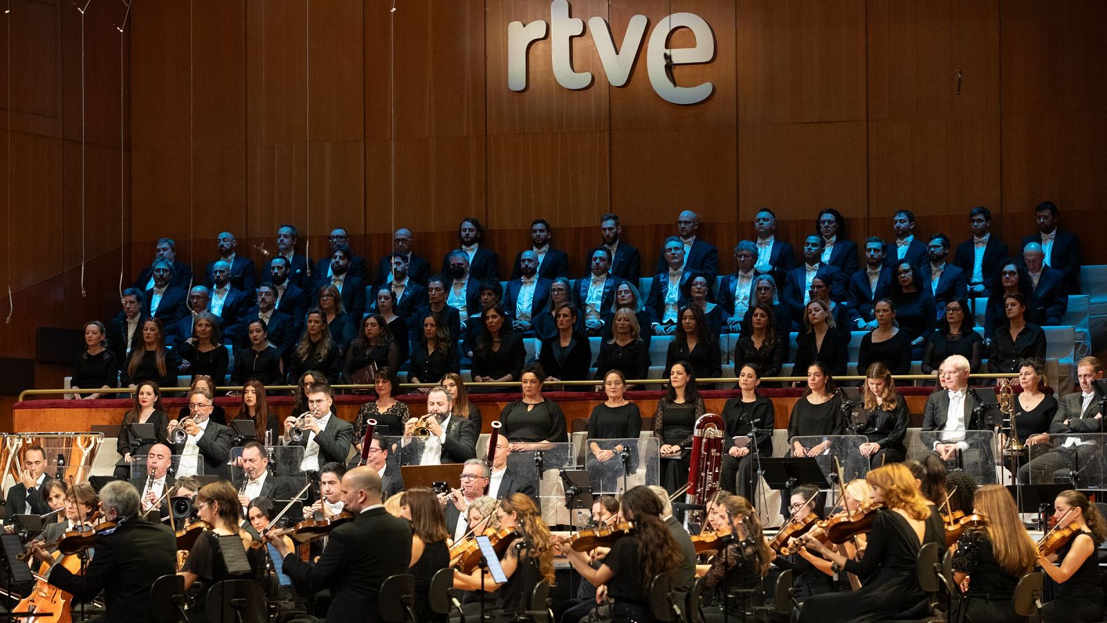 Orquesta y coro interpretando un concierto navideño. Músicos con atuendos oscuros tocando instrumentos de cuerda, viento y percusión. Coro situado en grada superior.