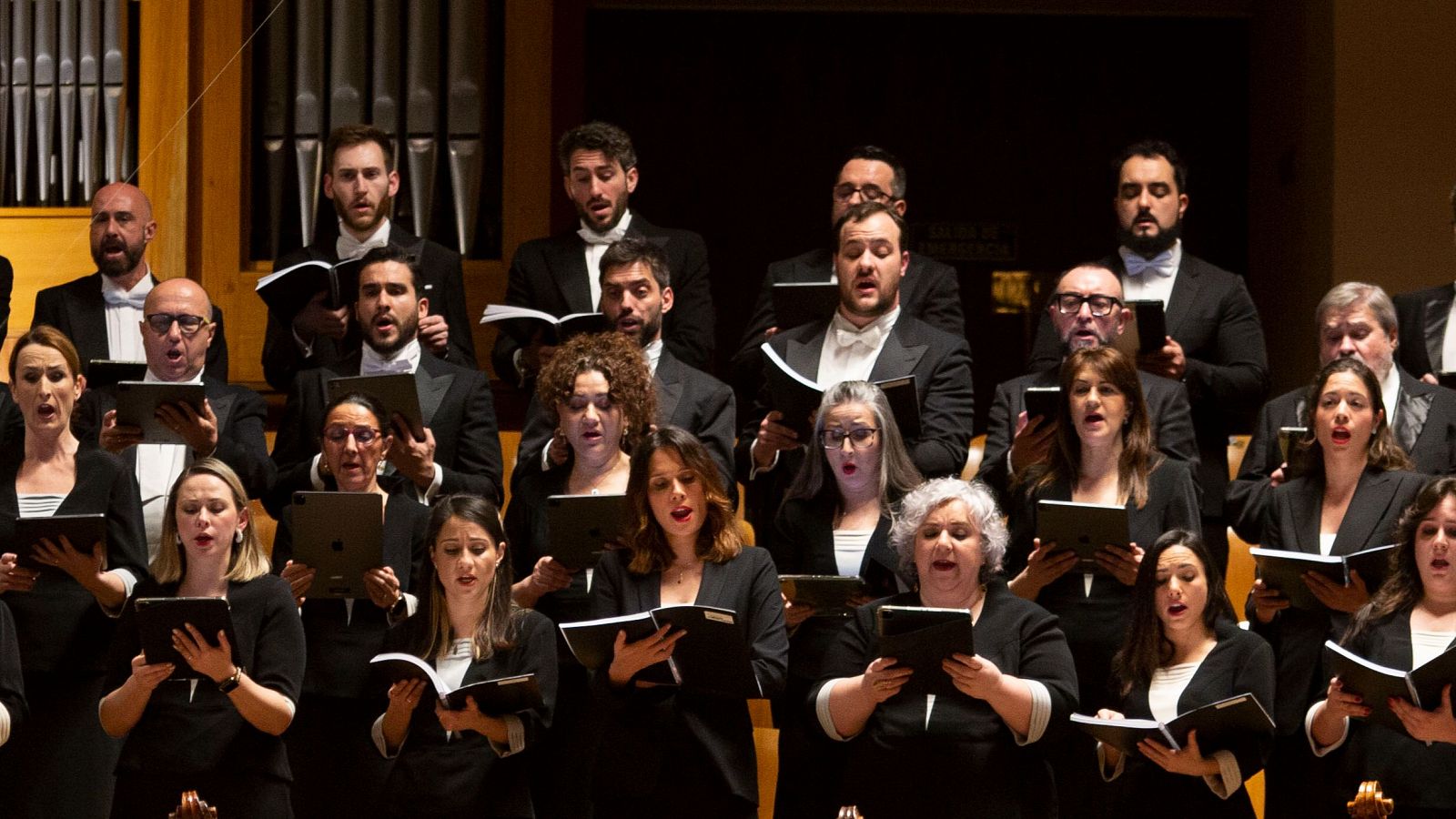 Actuación del Coro RTVE: numerosos cantantes, vestidos de negro, interpretan una pieza musical con partituras y tabletas.  Ambiente de concierto.