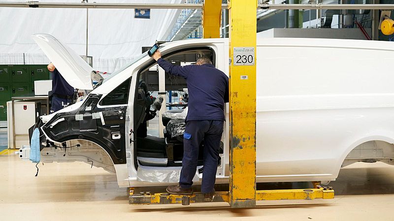 Trabajador en una fábrica ensamblando una furgoneta blanca sobre una plataforma elevadora.  Utiliza una tableta y se observa una etiqueta con el número 230.