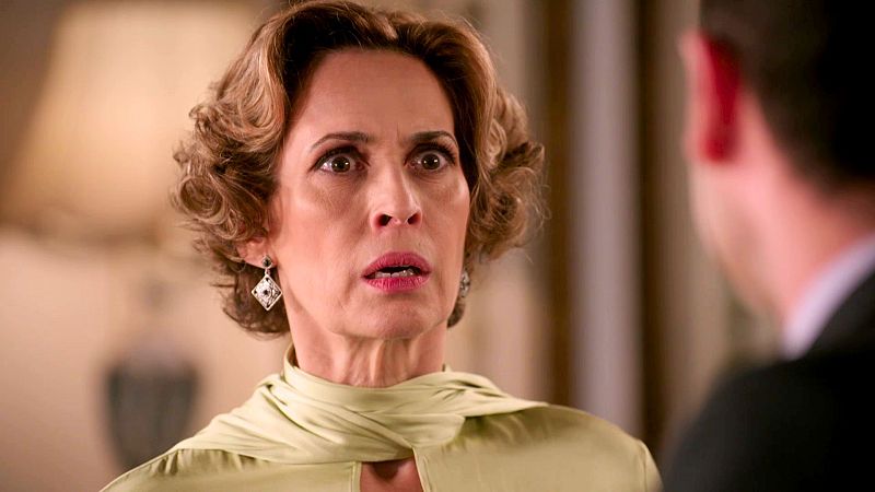 Mujer con blusa verde y pelo castaño rizado, expresión de sorpresa, habla con un hombre (solo visible parcialmente) en un interior cálido.