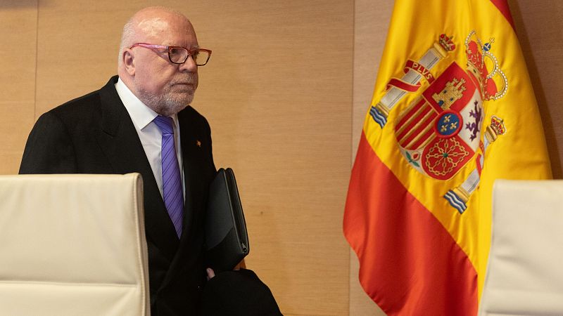 José Manuel Villarejo Pérez, ex comisario, aparece sentado, con traje oscuro, camisa blanca, corbata morada y carpeta negra.  Fondo con bandera de España.