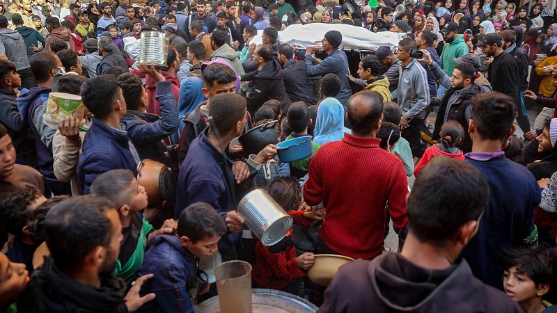Funeral en el que una multitud rodea un cuerpo cubierto con una tela blanca.  Presentes recipientes metálicos y de plástico.
