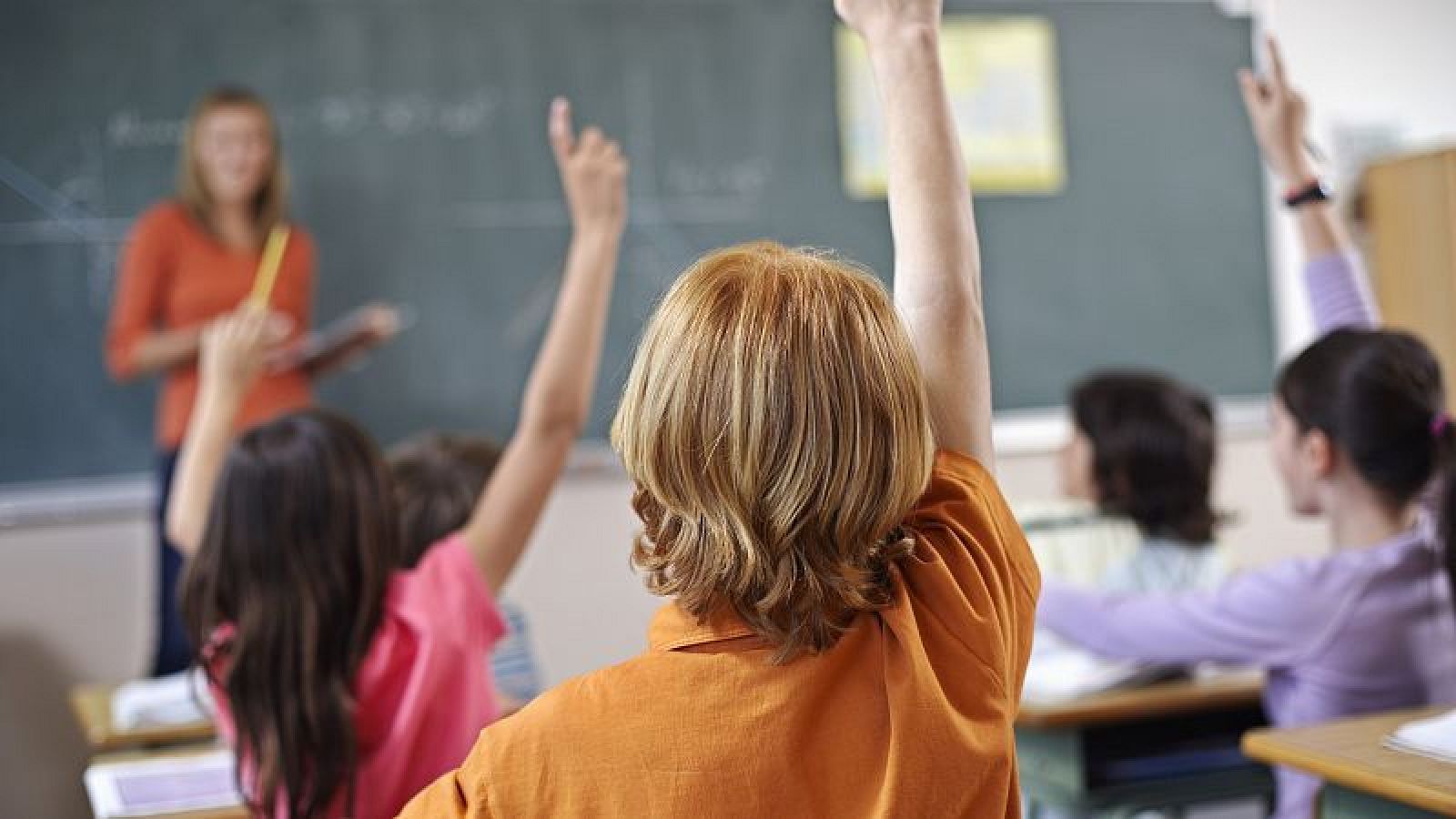 Escena de clase participativa: profesora con camisa naranja explica lección en pizarra, alumnos con las manos levantadas.