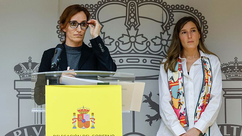 Ministra en atril presentando proyecto de salud mental, con gafas y traje oscuro; acompañada por otra mujer con camisa blanca y pañuelo.  Escudo de España visible.