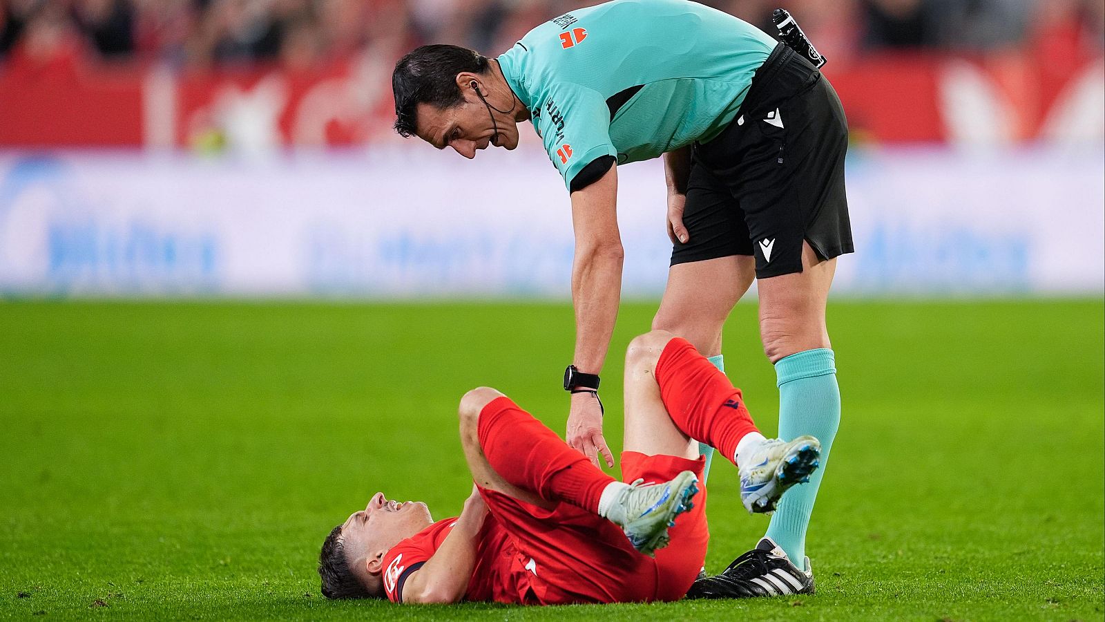 Jugador de fútbol lesionado en el césped. Un árbitro se acerca para asistirle. Se observa el campo de juego y parte de las gradas.