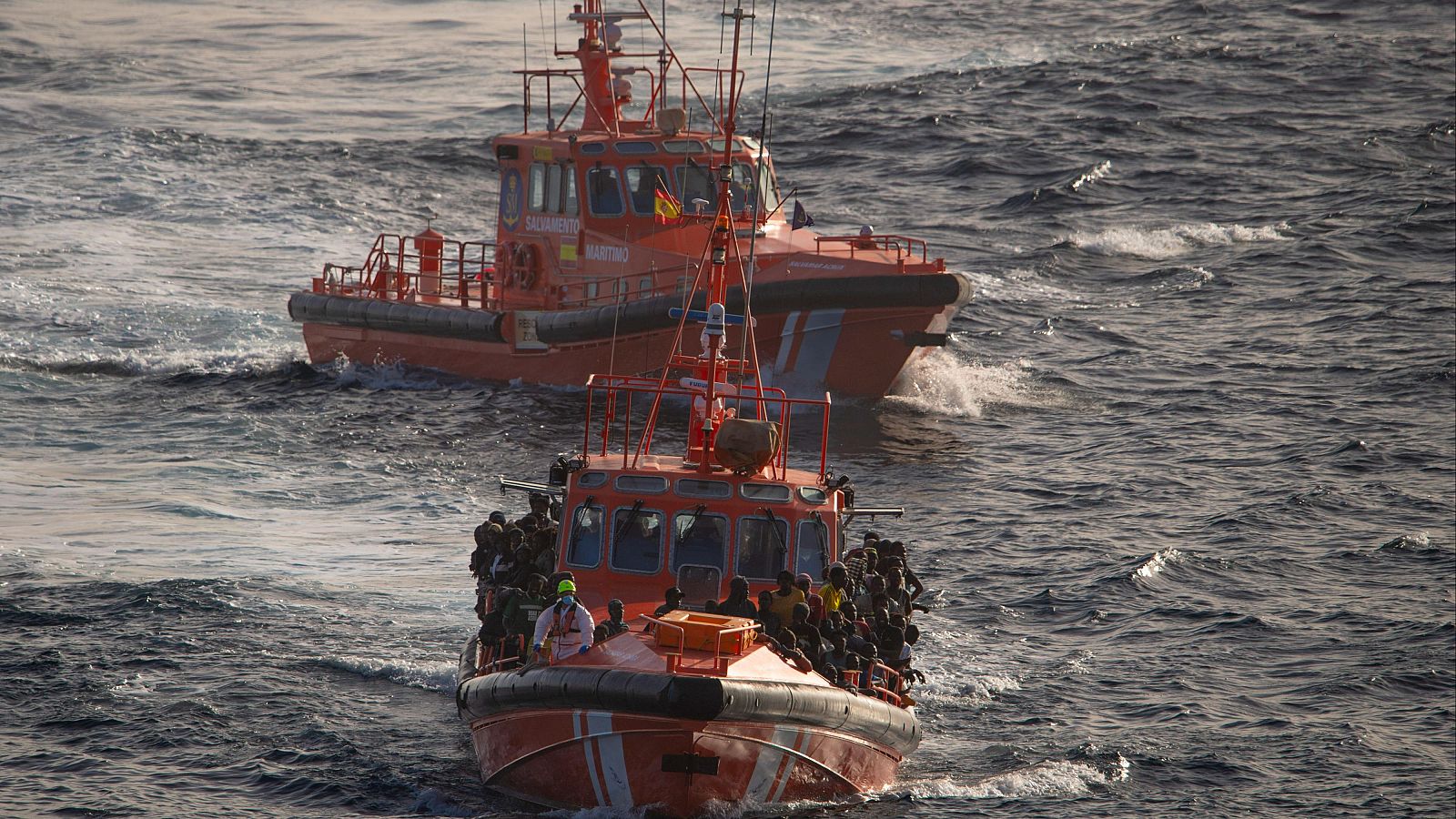 Rescate marítimo: Dos lanchas, una con numerosos migrantes rescatados y otra con bandera española, en mar con pequeñas olas.  Un rescatador con chaleco claro está entre los migrantes.