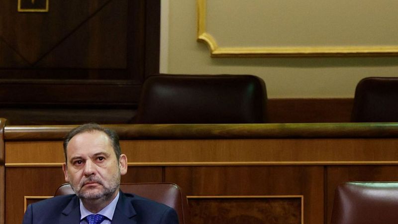 Exministro español sentado en una sala con mobiliario de madera y detalles dorados. Lleva traje oscuro, camisa blanca y corbata azul, con expresión seria.