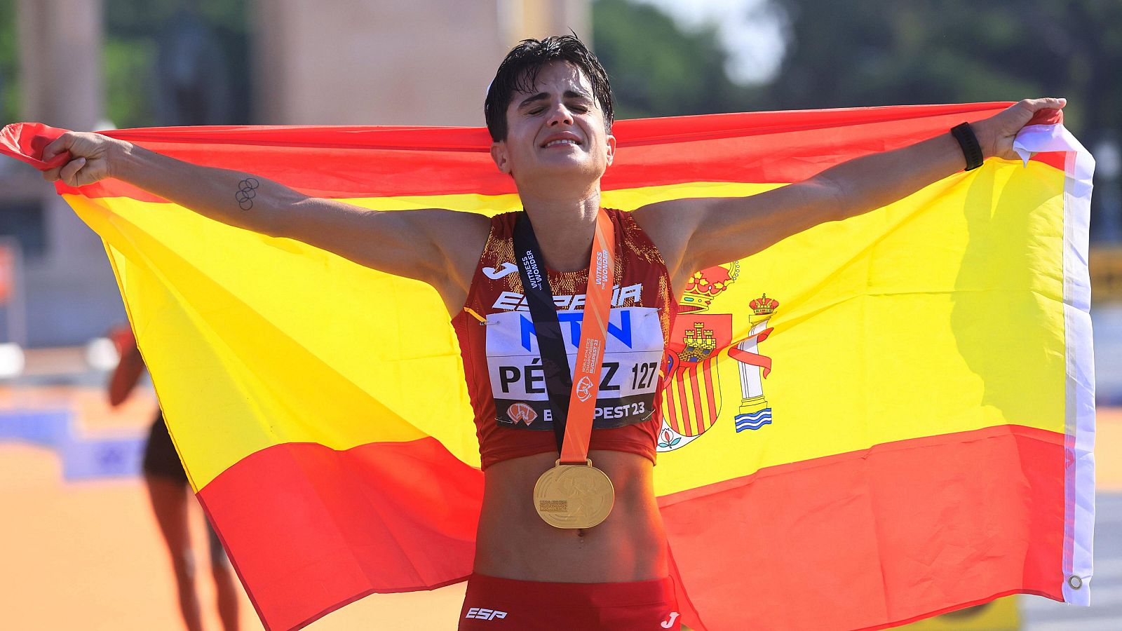 Atleta con uniforme rojo y medalla de oro celebra victoria, mostrando bandera española y tatuaje de anillos olímpicos.