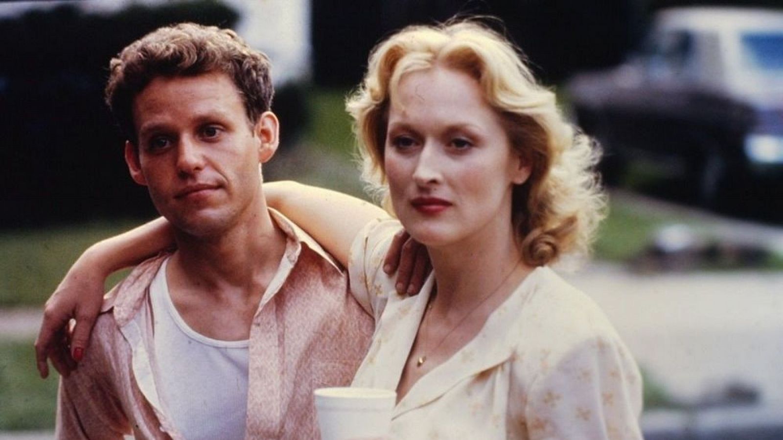 Meryl Streep, con vestido claro y collar, y un hombre con camisa de manga corta, en escena exterior.  Vaso en primer plano.