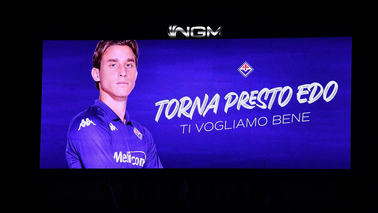 Pantalla gigante con foto de futbolista en camiseta morada con logo Kappa y patrocinador "Melícom". Mensaje: "TORNA PRESTO EDO, TI VOGLIAMO BENE".