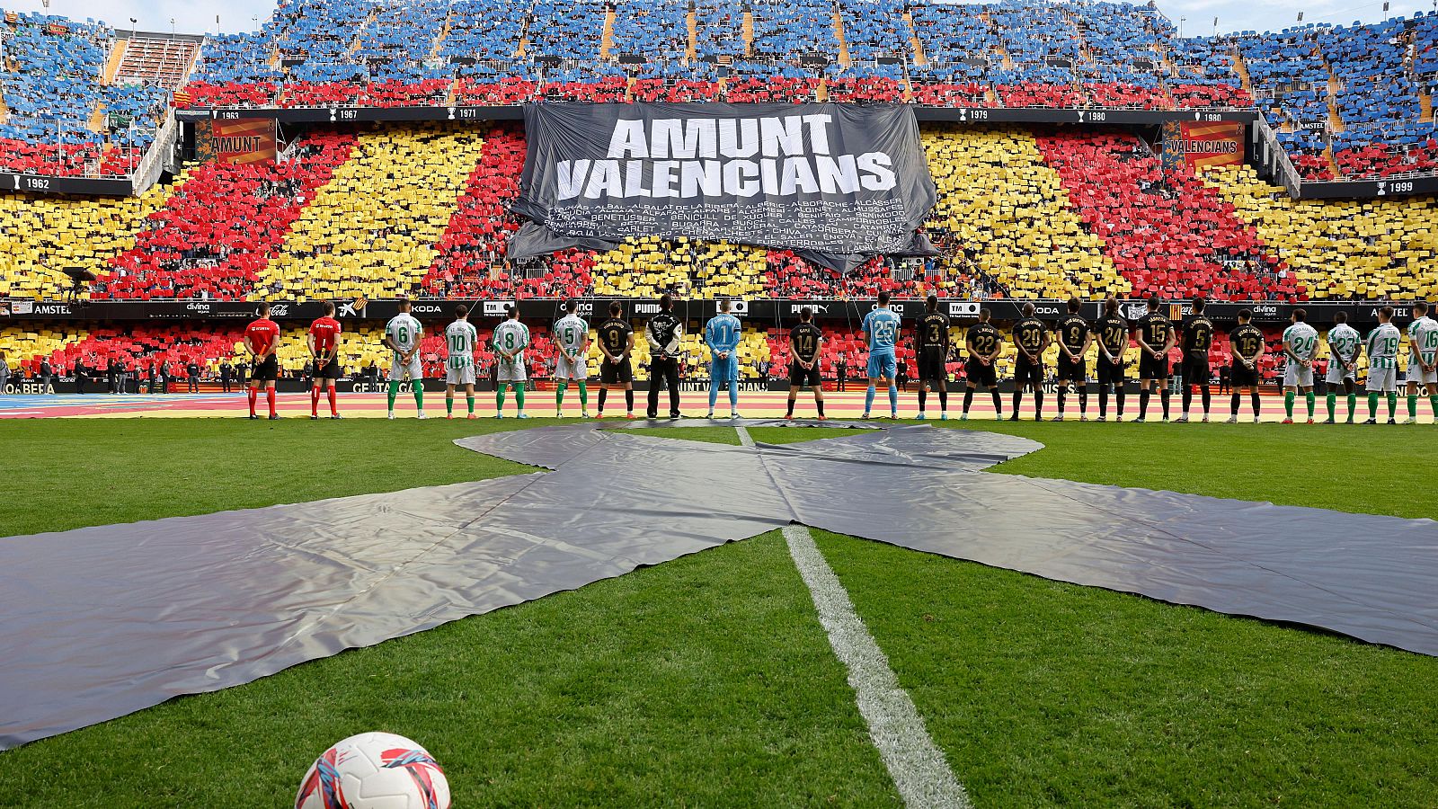 Minuto de silencio en un estadio de fútbol. Jugadores de dos equipos con uniformes distintos y árbitros alineados en el centro del campo. Pancarta visible en las gradas.