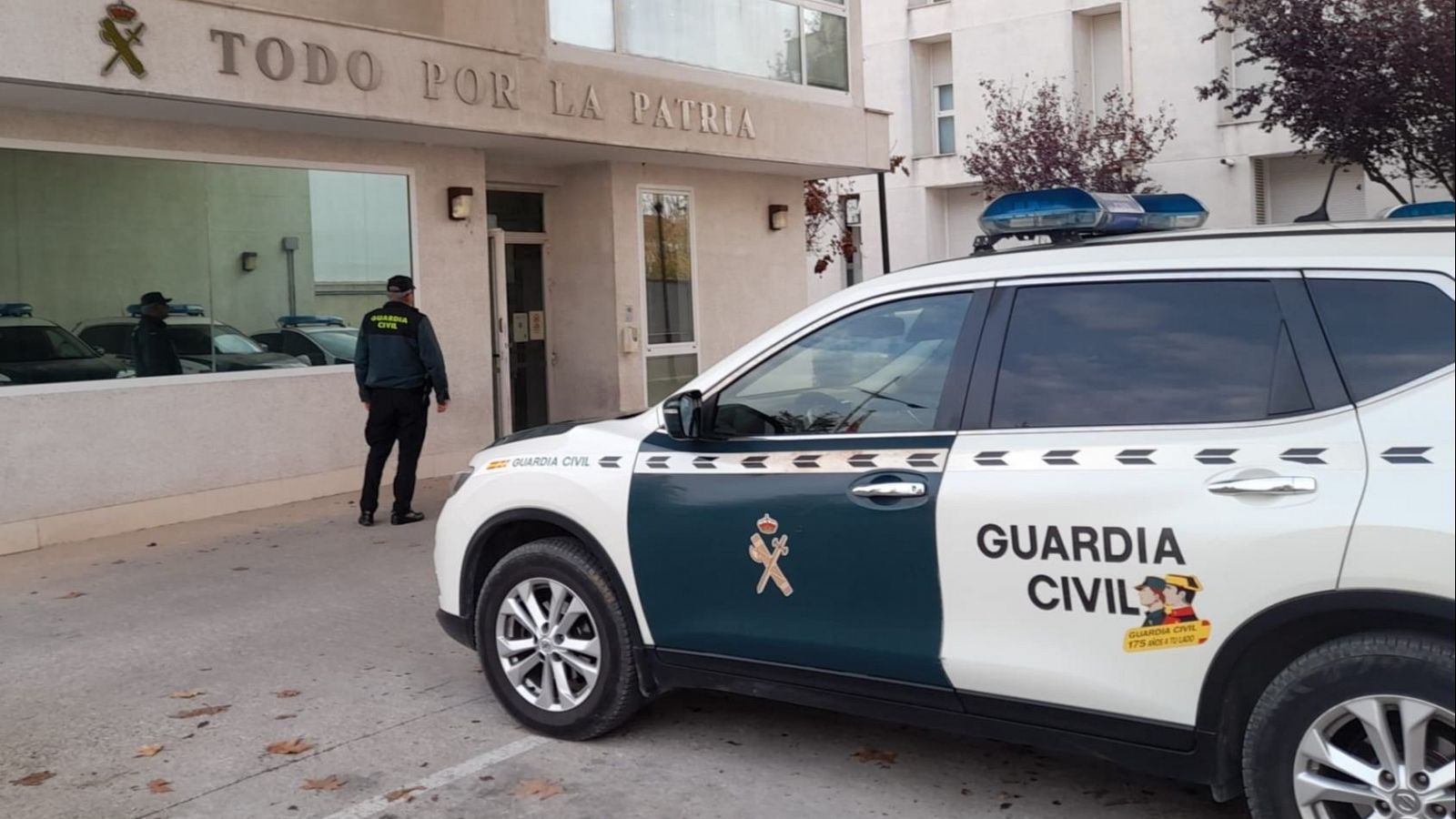 Edificio de dos plantas con la inscripción "TODO POR LA PATRIA", un coche patrulla blanco y verde aparcado delante y un agente de la Guardia Civil en la entrada.