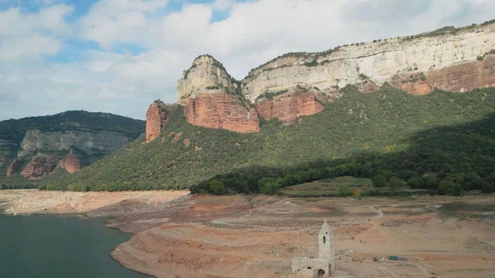 Vista panorámica del Pantà de Sau con bajo nivel de agua, mostrando tierra marrón y vegetación verde en las montañas.  Se aprecia una estructura antigua en la orilla.