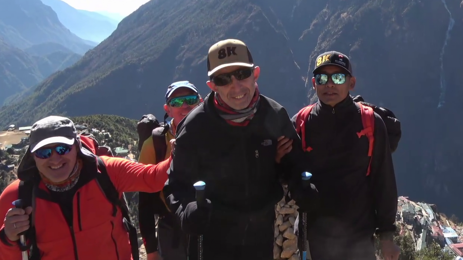 Expedición montañosa: cuatro personas con ropa de abrigo y bastones de trekking en una ladera, con un valle visible al fondo.  Un hombre lleva una gorra con las letras "8K".