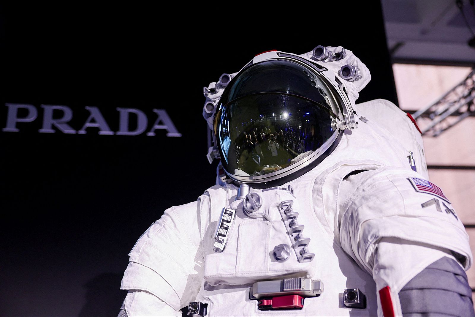 Traje espacial blanco con detalles en gris y rojo, bandera estadounidense y letras "AX" en el brazo. Visera reflectante. Diseño Prada y Axiom Space para Artemis III.