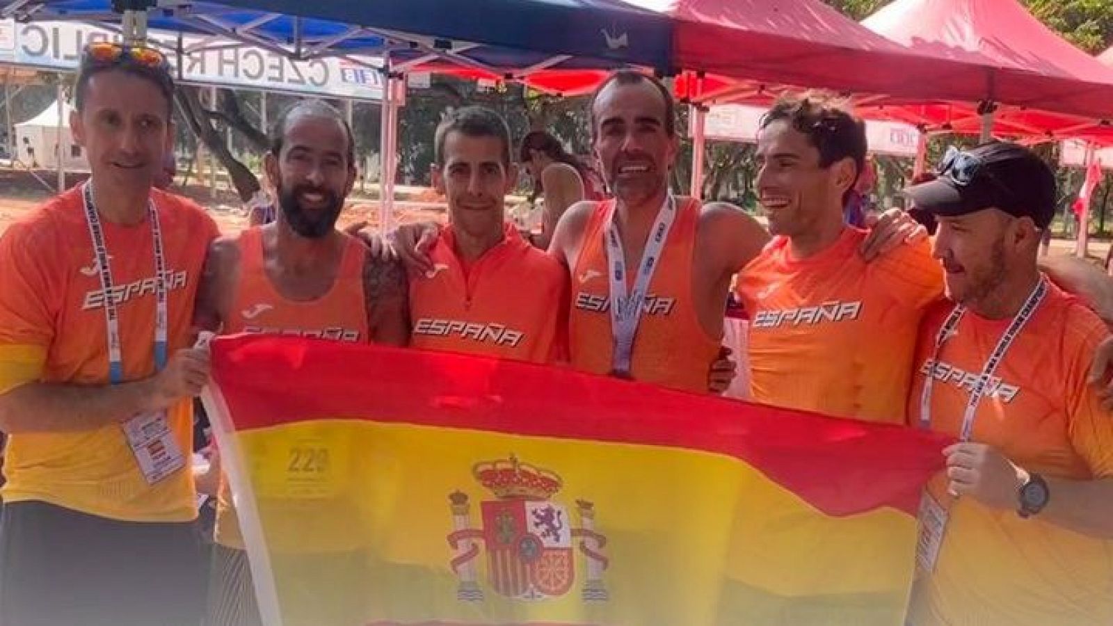 Atletas españoles, con camisetas naranjas y una medalla de plata, celebran su victoria en un campeonato al aire libre.  Sostienen una bandera española.