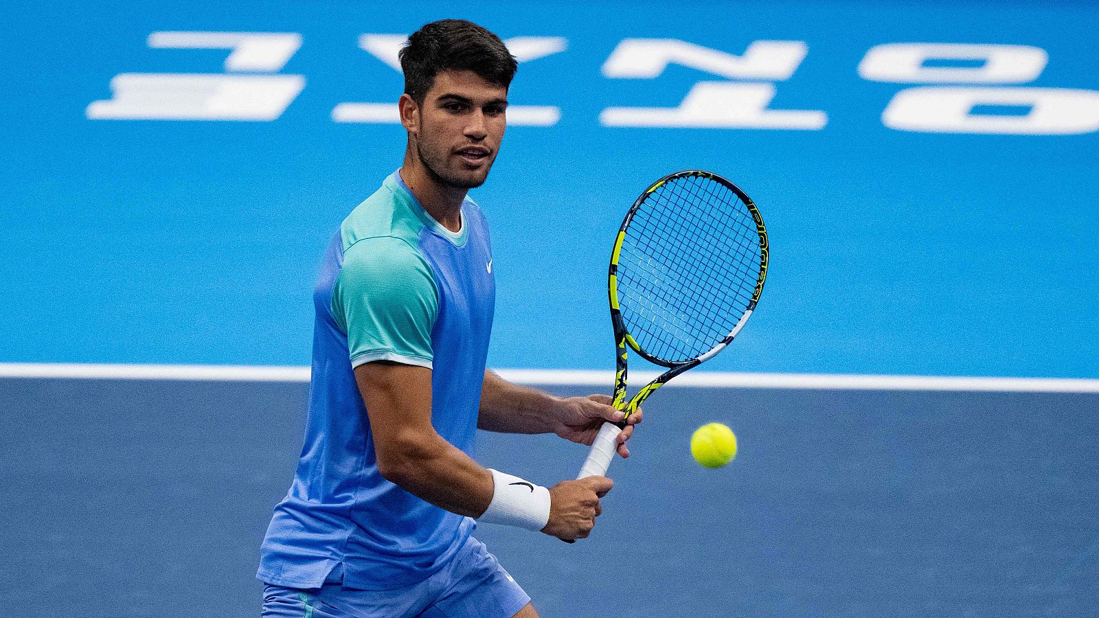 Tenista con camiseta azul y pantalón corto golpeando una pelota amarilla. Lleva una muñequera y una raqueta. Fondo con parte del logo del torneo.