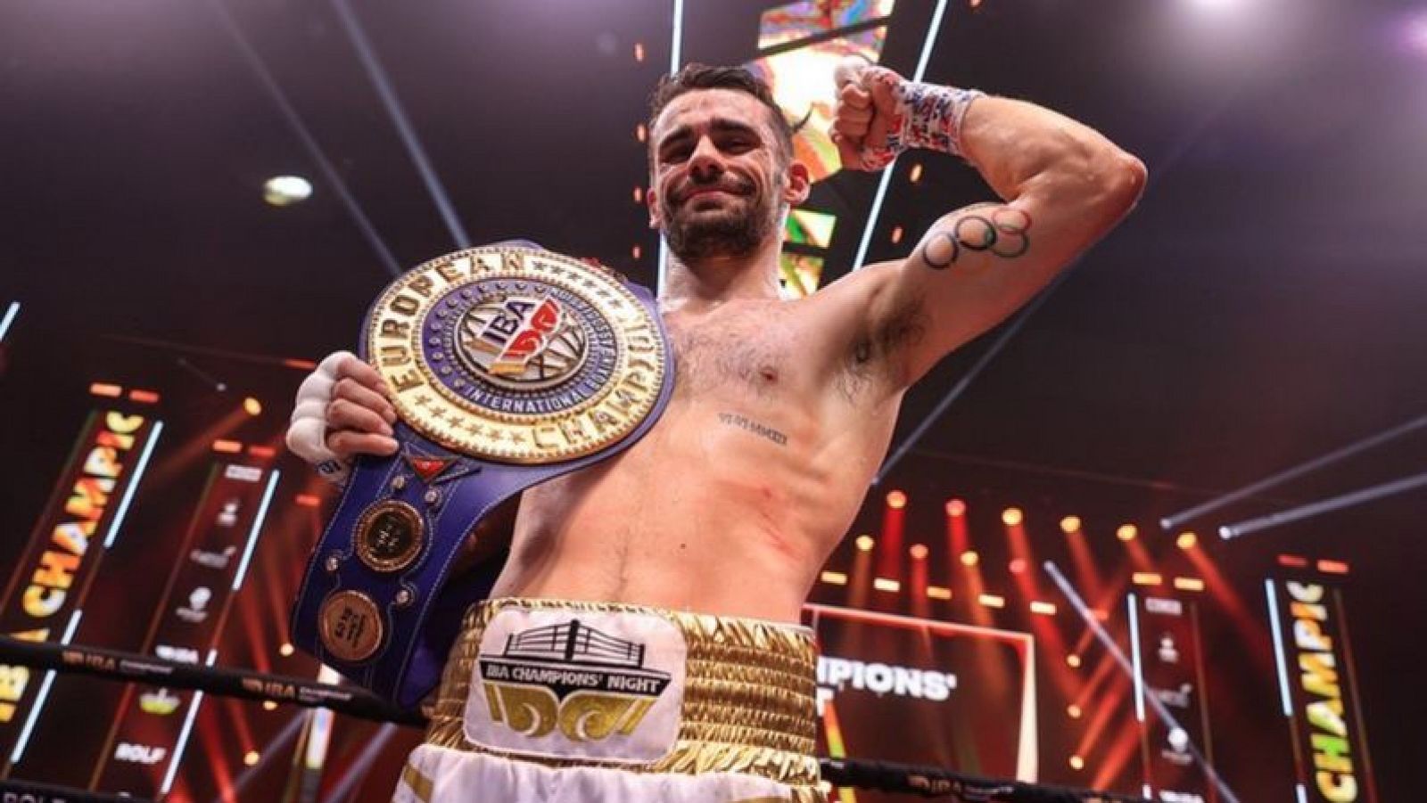 Boxeador celebrando victoria, mostrando tatuaje de anillos olímpicos y cinturón de campeón azul con detalles dorados que indican 'EUROPEAN', 'IBA', 'INTERNATIONAL' y 'CHAMPION'.