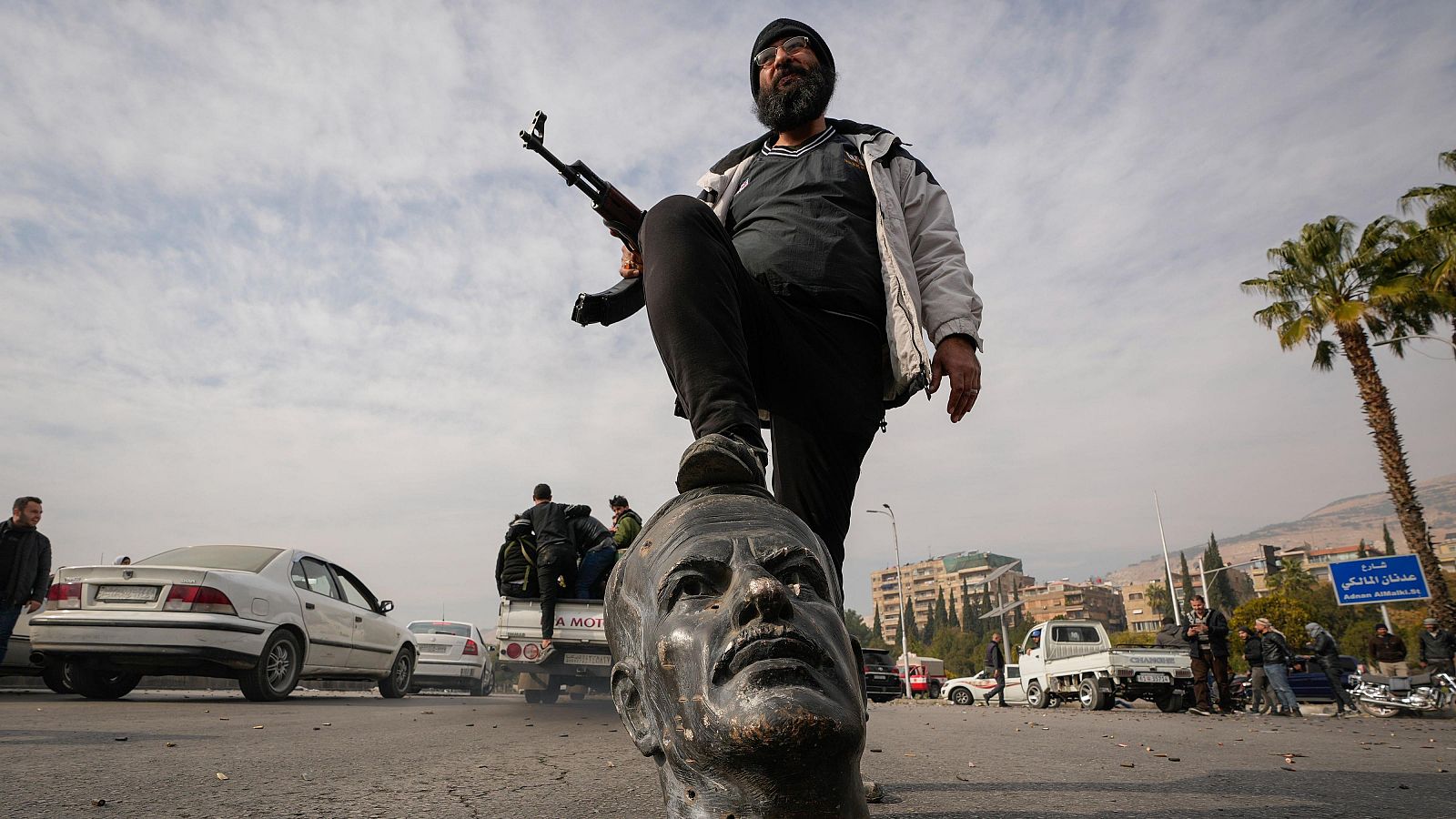 Hombre con rifle AK-47 sobre estatua derribada en un contexto de conflicto.  Se observan otros individuos y vehículos en el entorno.