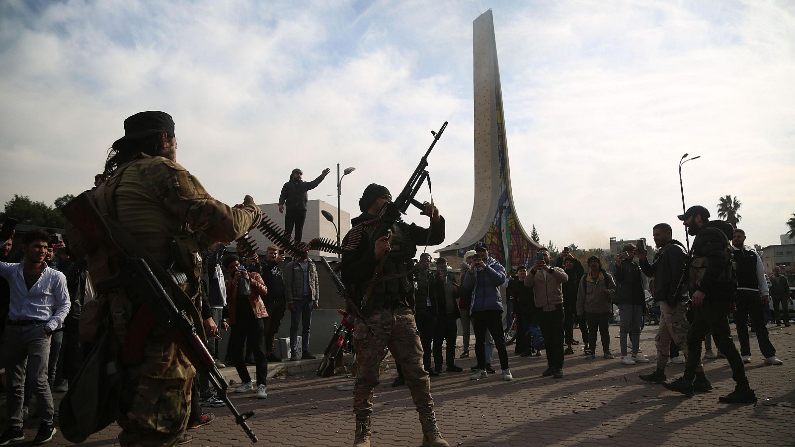 Enfrentamiento en Siria: dos militares armados, un civil sobre un muro y una multitud observando y grabando con móviles.