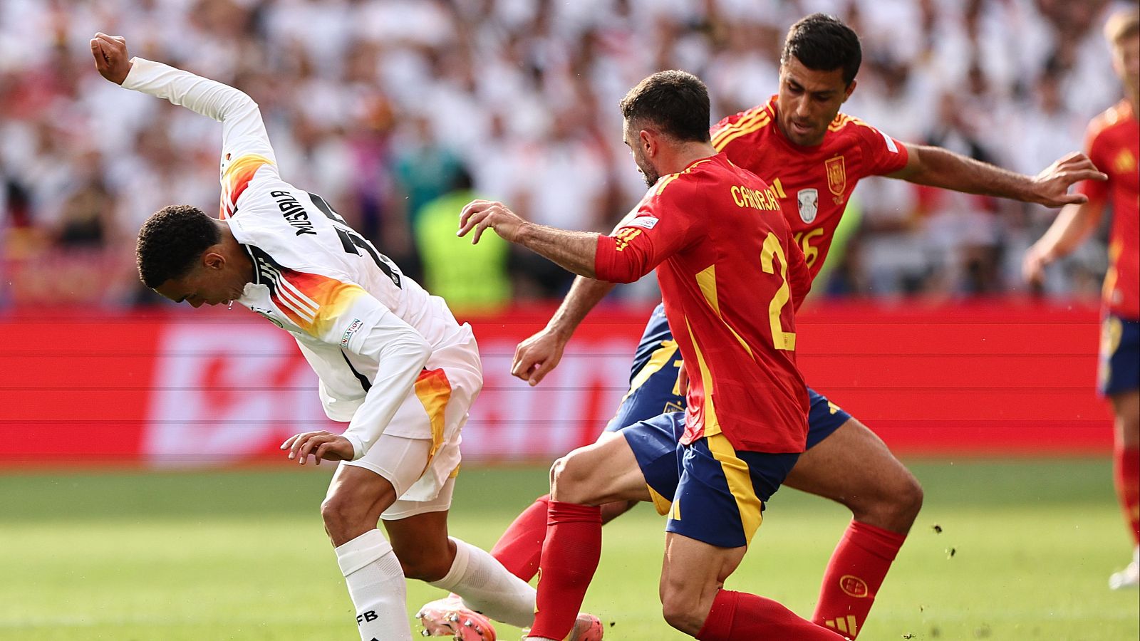 Carvajal (dorsal 12) defiende contra un jugador rival con camiseta blanca, naranja y amarilla que parece desequilibrarse.  Intensa acción en un partido de fútbol.