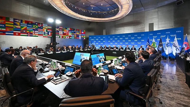 Reunión en sala moderna con asistentes alrededor de una mesa en forma de U, cada puesto con micrófono y pantalla;  panel con logo FIFA y banderas en el fondo.
