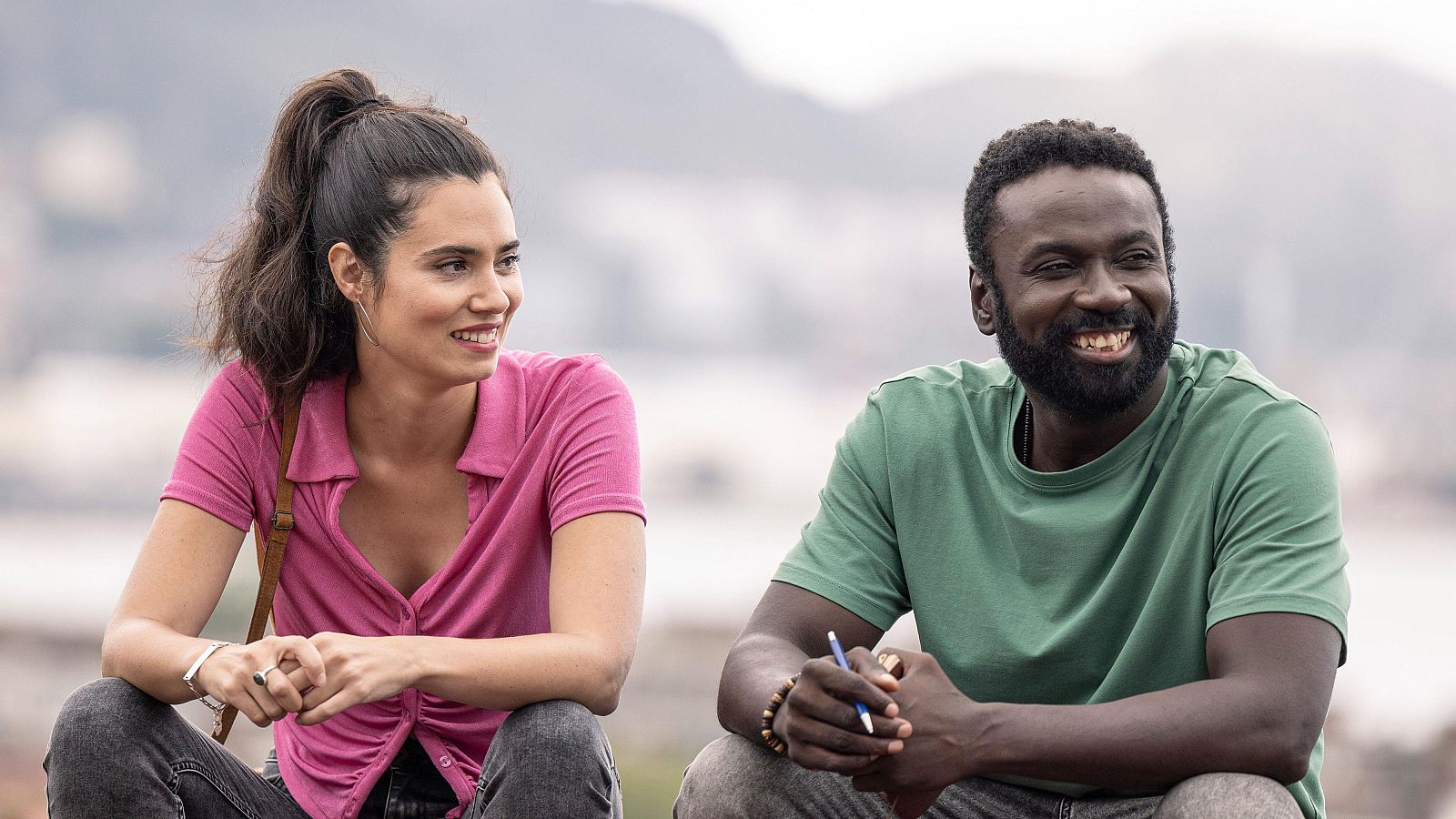 Dos actores, hombre con camiseta verde oliva y mujer con camiseta rosa, conversan sonrientes en un entorno exterior.  Posible escena de una serie de televisión.