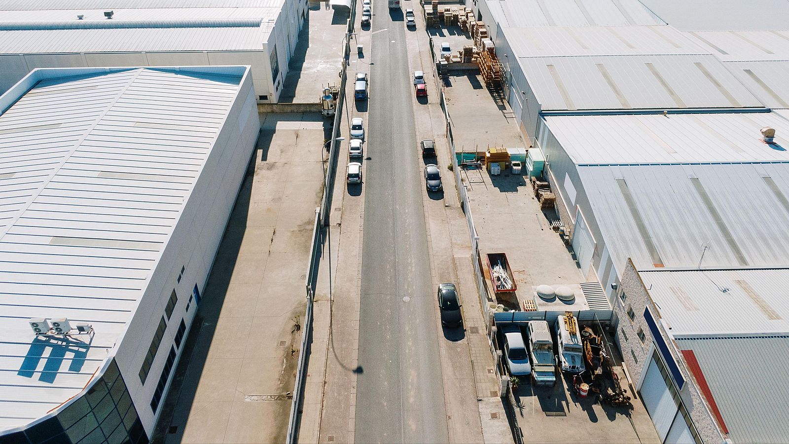 Vista aérea de una calle con naves industriales a ambos lados, coches y furgonetas aparcados, y materiales de construcción visibles.