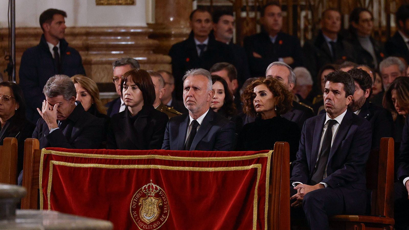 Funeral solemne con figuras políticas, entre ellas Carlos Mazón con gesto de tristeza, y otras personas vestidas de luto. Ambiente de solemnidad en un espacio con arquitectura religiosa.