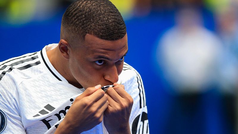 Primer plano de Kylian Mbappé besando su camiseta blanca con detalles negros y las tres rayas de Adidas. Fondo azul difuminado.